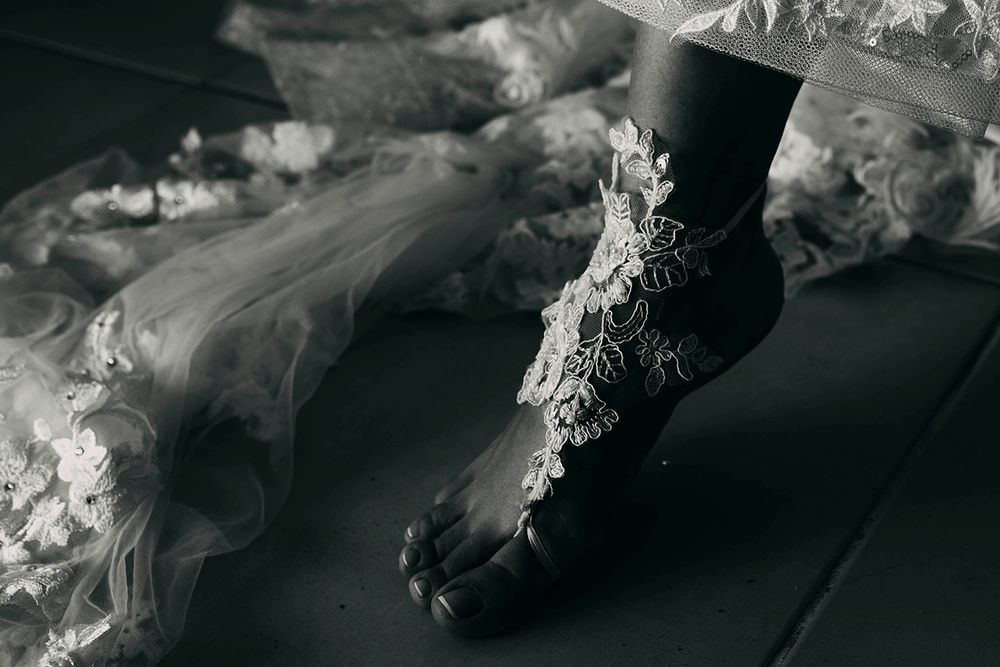 Close-up black and white photo of bride's foot wearing delicate lace floral anklet on dark floor.