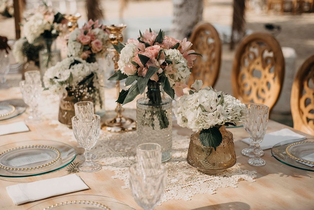 Elegant wedding reception table with floral centerpieces, crystal glasses, lace runner, and gold vases holding white and pink blooms.