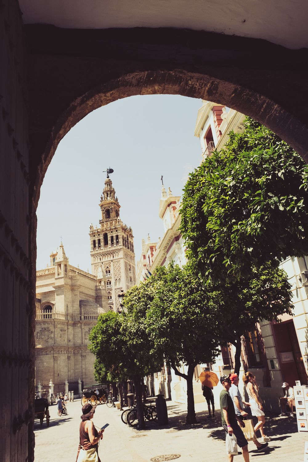 Giralda tower viewed through a stone arch with orange trees and tourists in Seville, Spain.