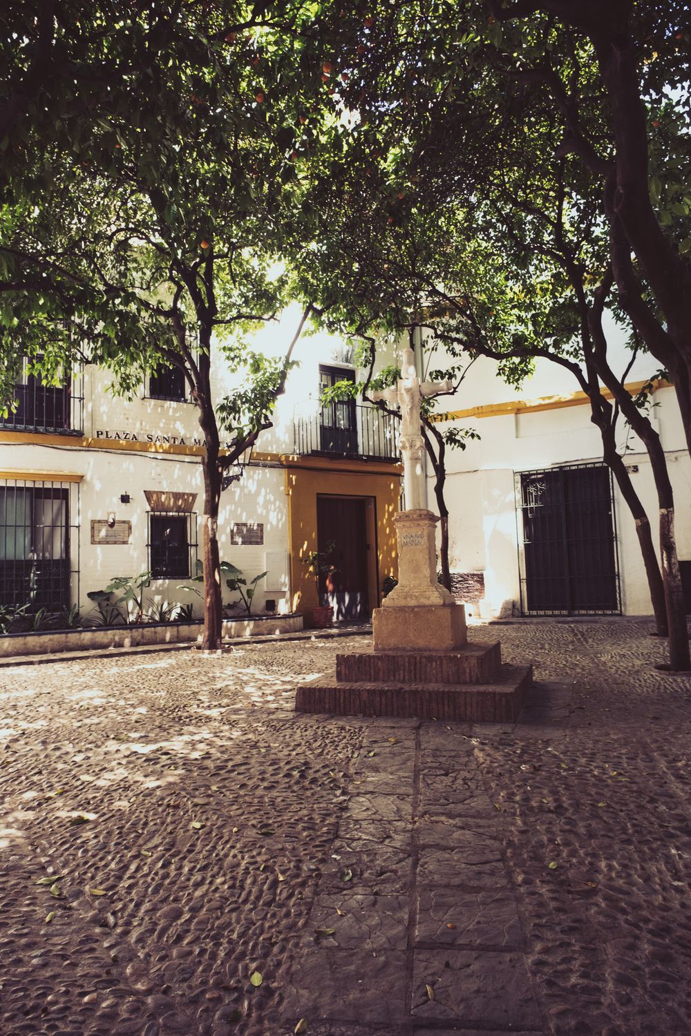 Cobblestone plaza with a stone cross monument shaded by trees in historic Seville, Spain.