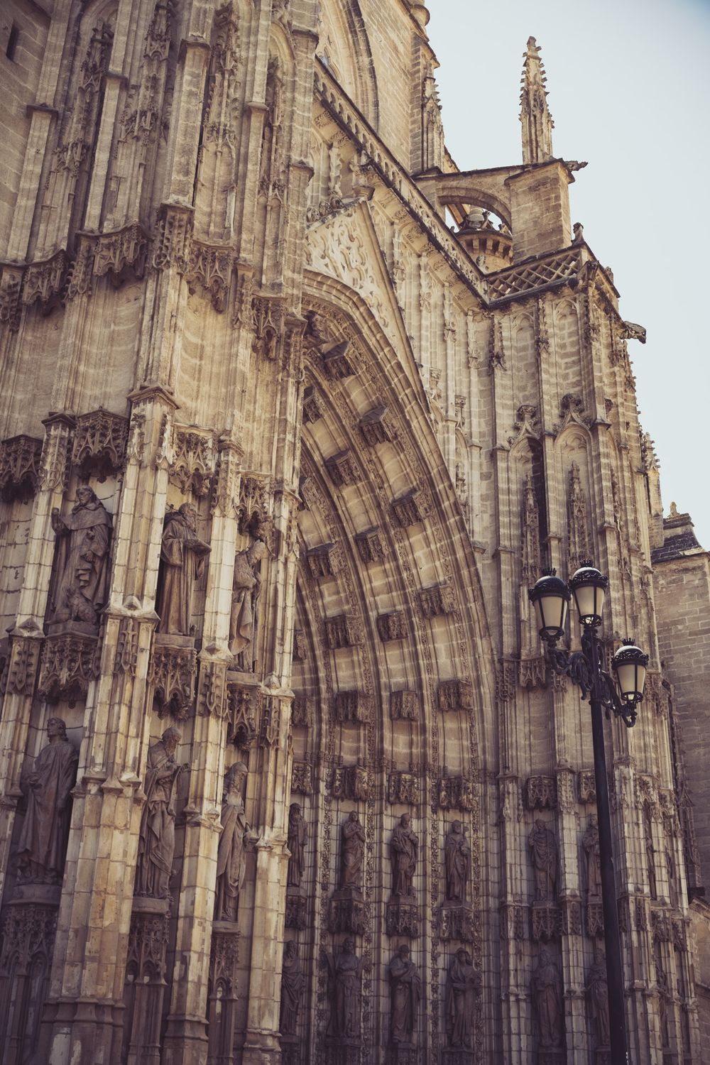 Gothic cathedral exterior detail showing intricate stone carvings, pointed arches, and sculptural decorations with a vintage street lamp.