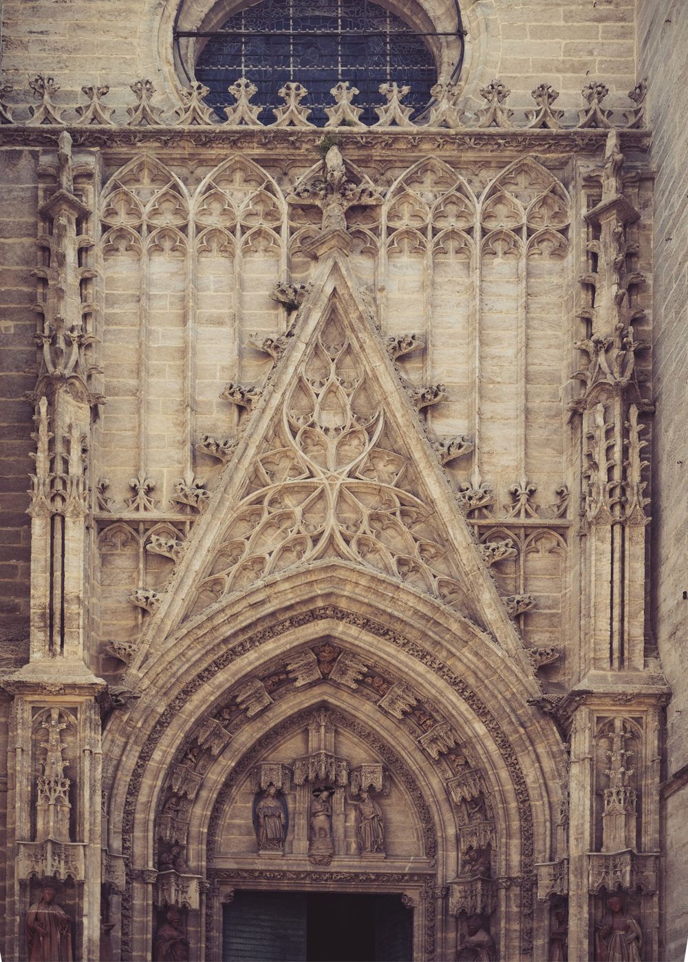 Gothic portal of Seville Cathedral featuring intricate stone carvings, pointed arch doorway, and ornate medieval religious sculptures.