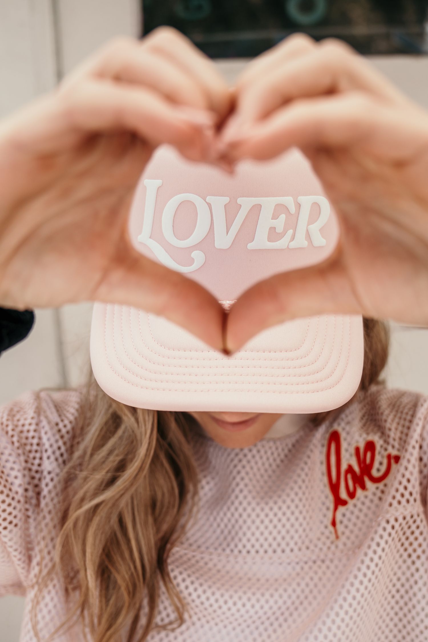 Woman making heart shape with hands over pink Lover trucker hat, wearing love-script mesh top.