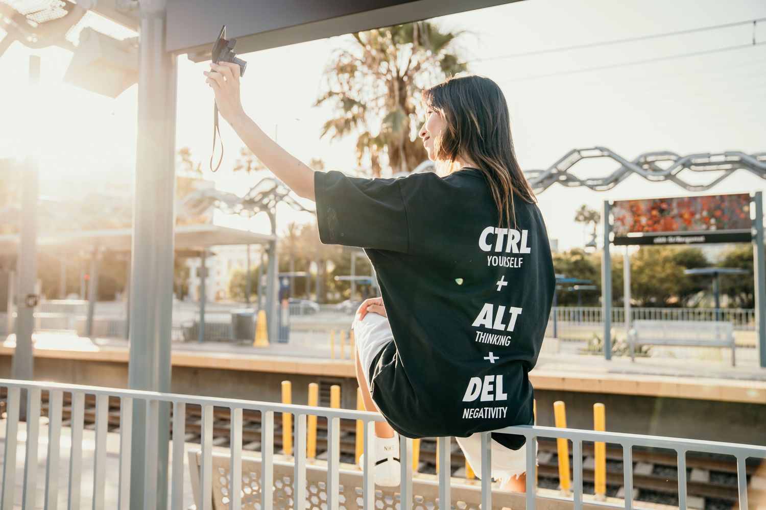Woman wearing black graphic tee with CTRL ALT DEL print, posing outdoors at sunny urban transit stop.