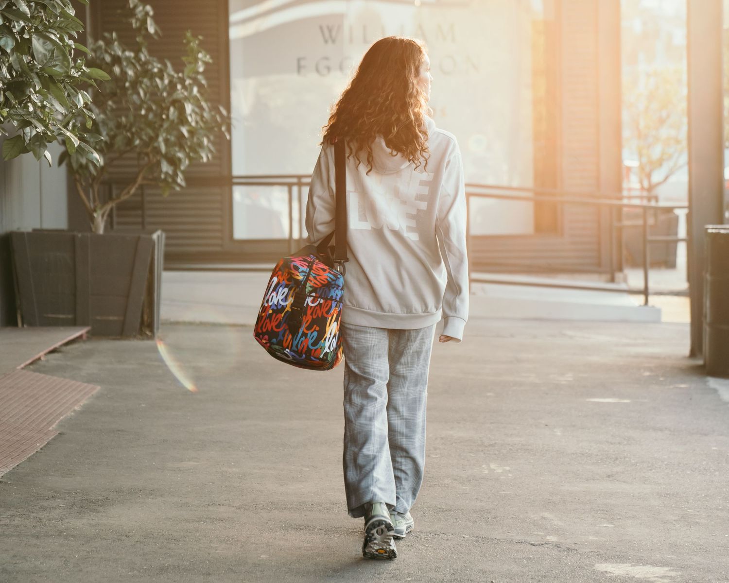 Young woman walking away carrying a colorful floral bag, wearing a white hoodie and gray jeans in urban setting.