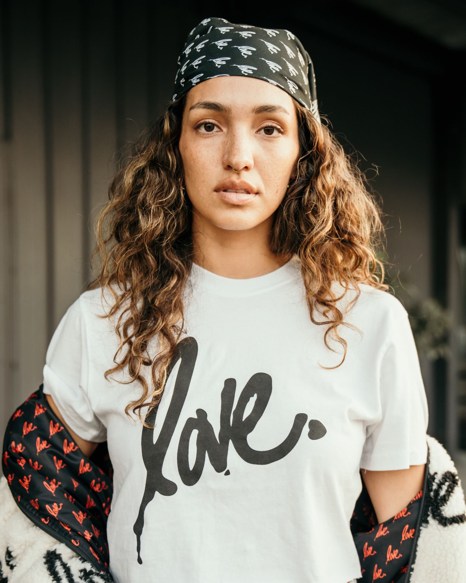 Young woman with curly hair wearing a black bandana and white Love graphic tee, posing outdoors.