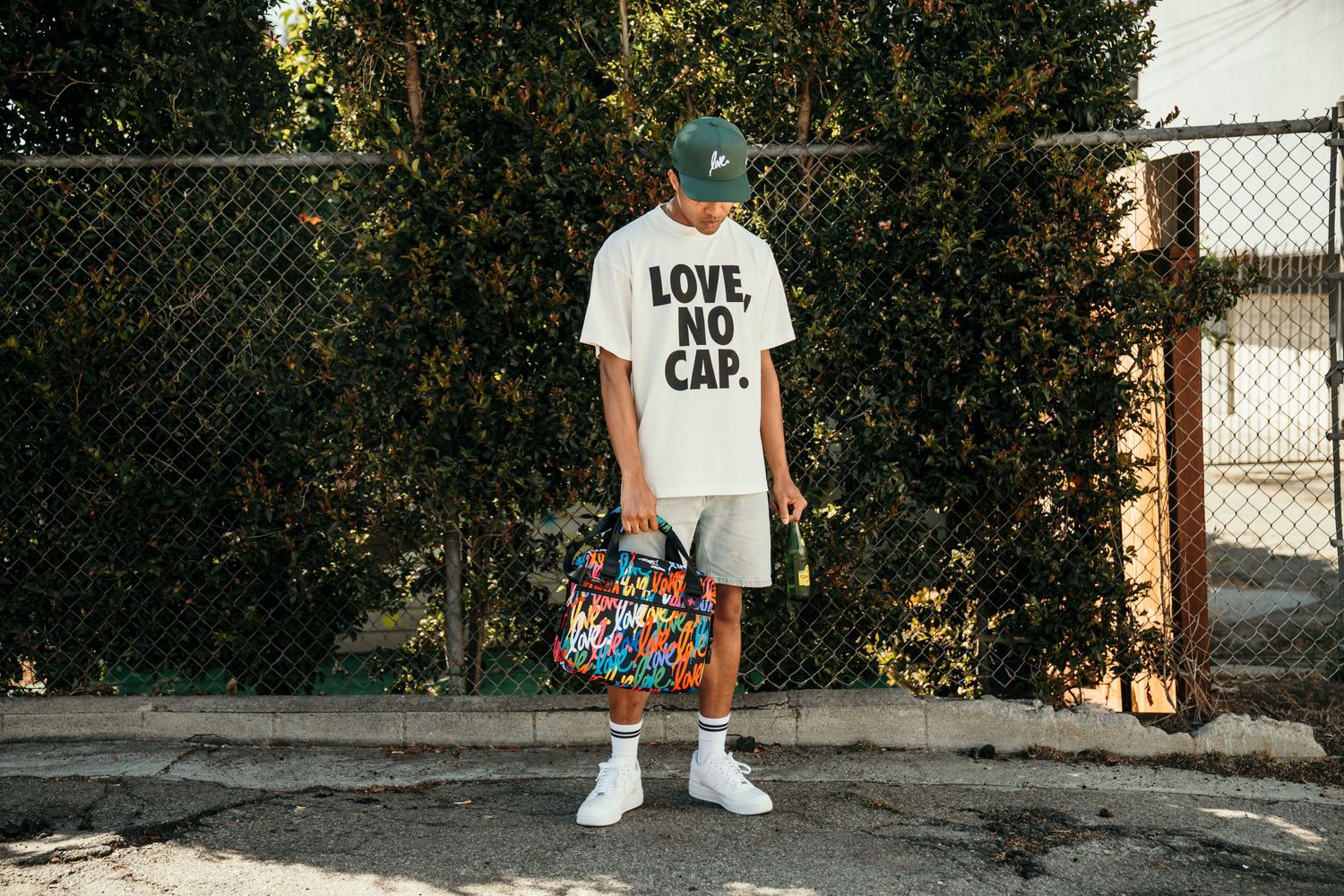 Man wearing white Love No Cap t-shirt, grey shorts, and white sneakers, holding colorful bag outdoors.