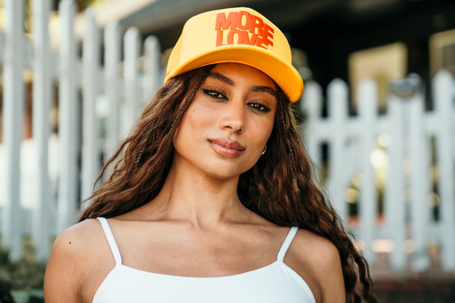 Young woman wearing an orange trucker hat and white tank top posing outdoors near a white fence.