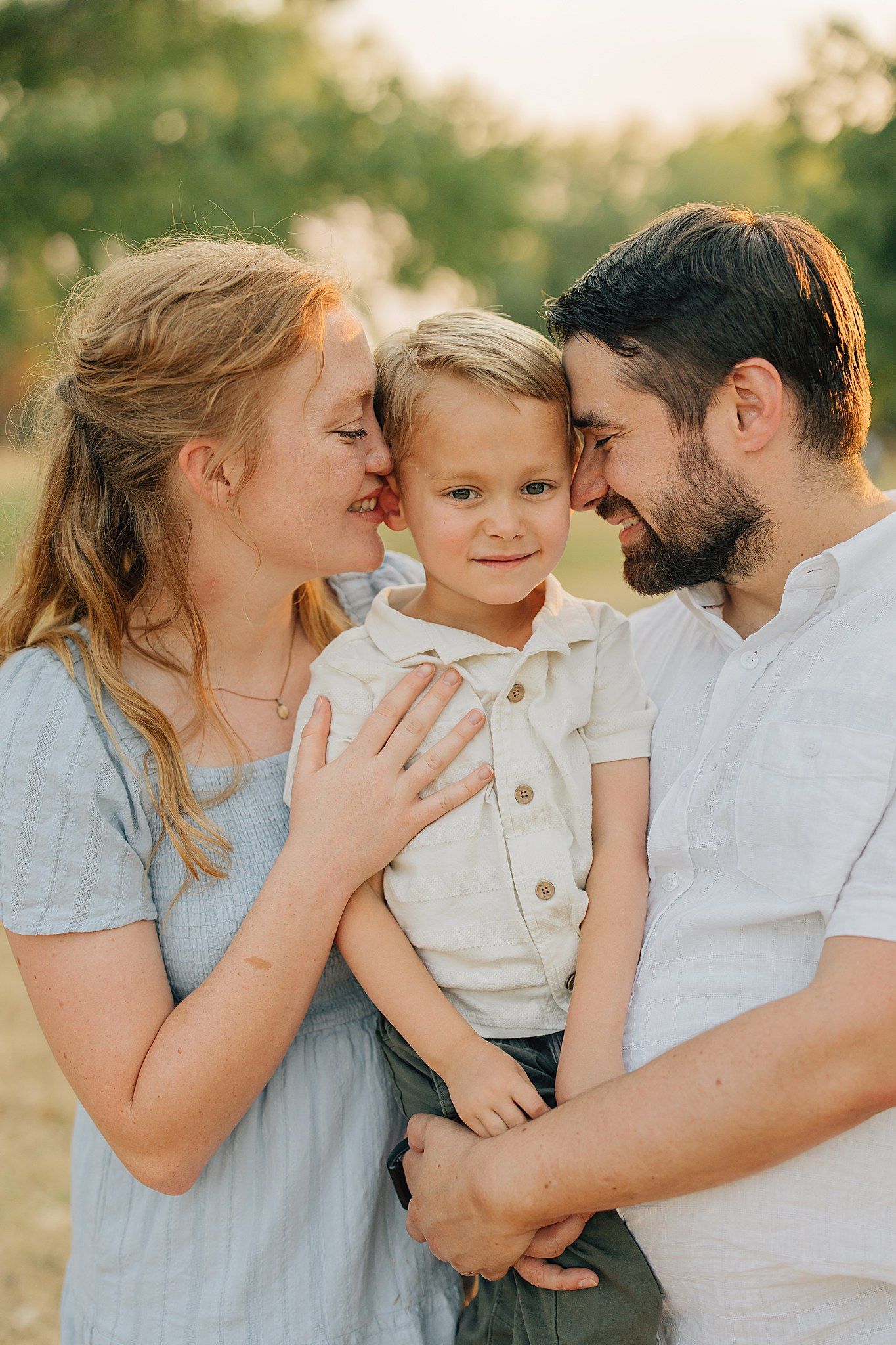 A joyful family portrait of parents lovingly embracing their young blond son outdoors in warm golden light.