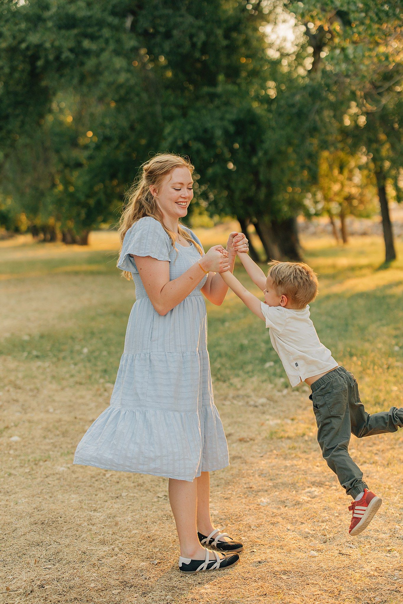 A smiling mother in a blue dress twirls her young son in a sunlit park.
