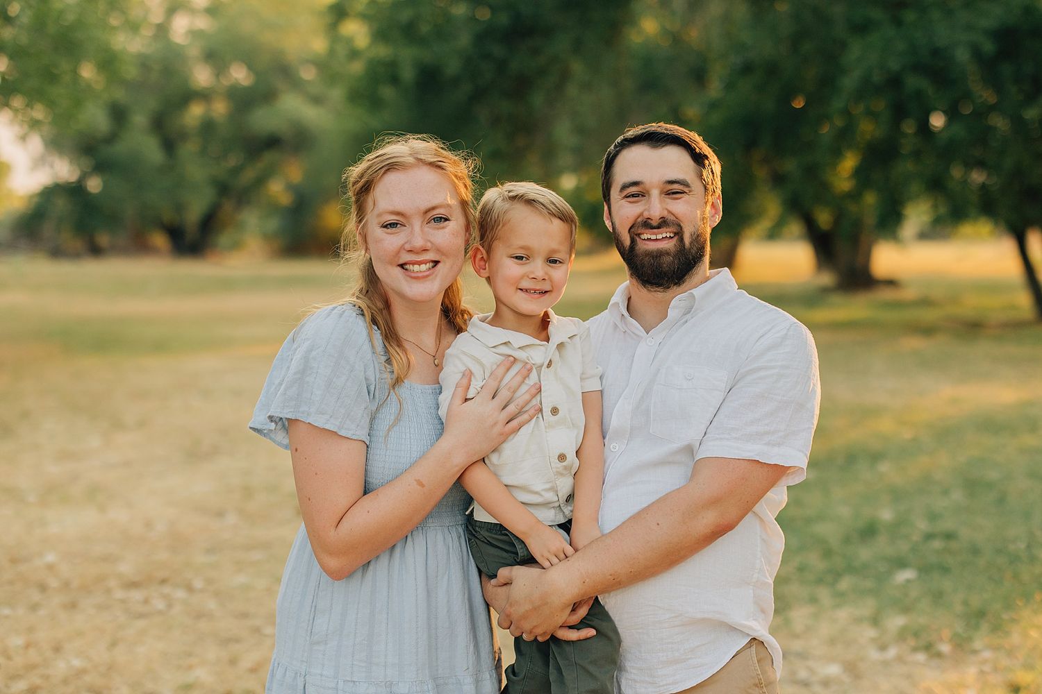 Happy family of three posing outdoors in a sunny park, parents holding young child, wearing casual summer outfits.