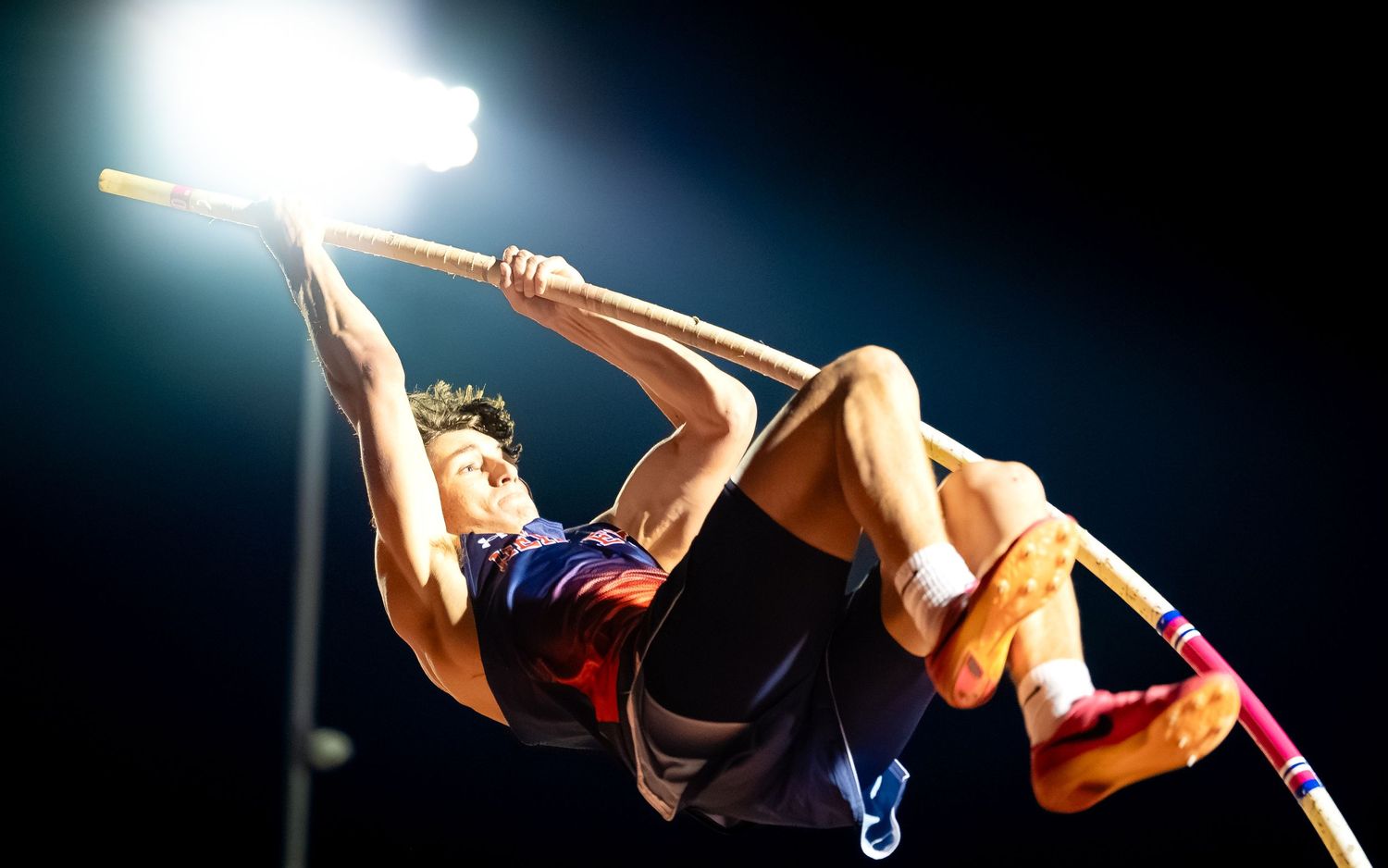 Hershey Trojans pole vaulter arching over the bar under the lights, Meet of Champions, by Deanna Fox, SportsFox Photography, Hershey PA