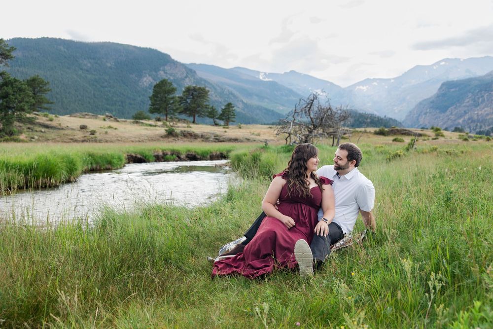Pregnant couple sitting in green meadow by a stream with Rocky Mountain peaks in background, woman in burgundy dress.
