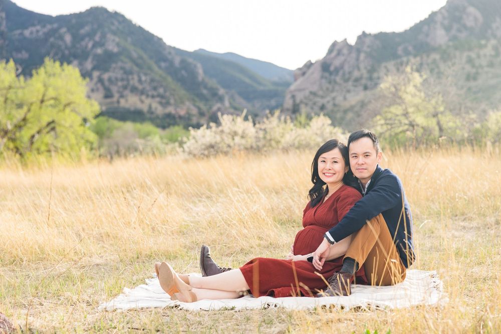 Couple sitting on blanket in golden field with Rocky Mountain backdrop during maternity photo session.