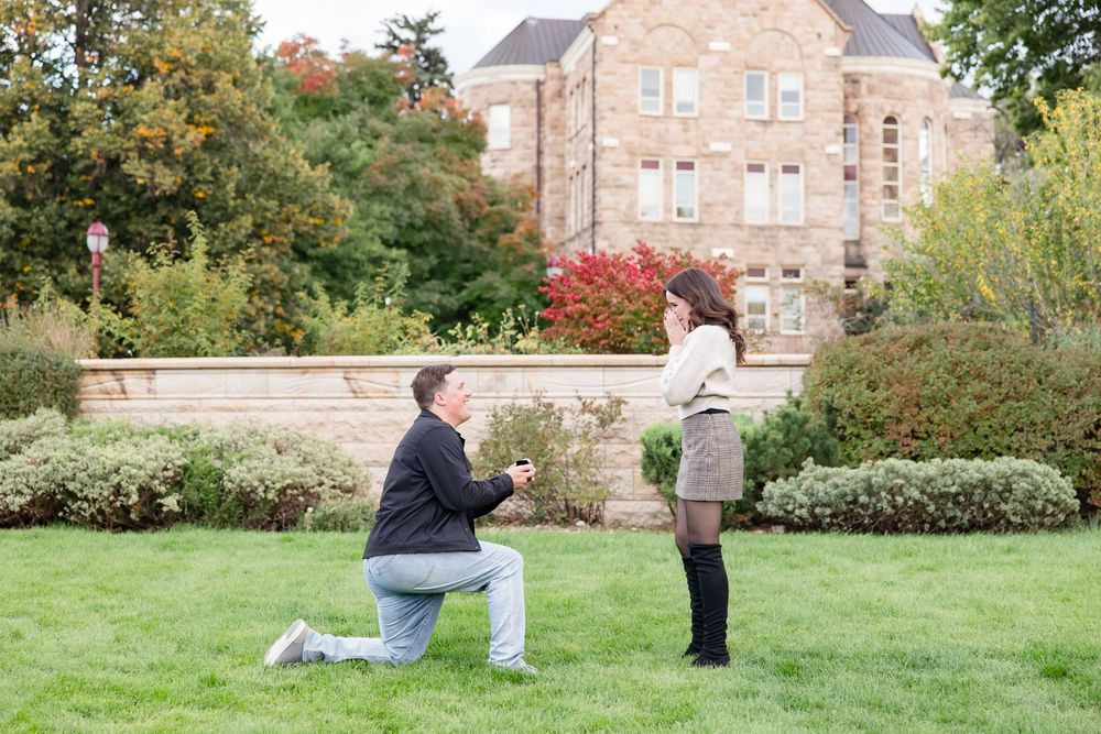 Man kneeling on one knee proposing to woman in lush garden with stone building backdrop during autumn.