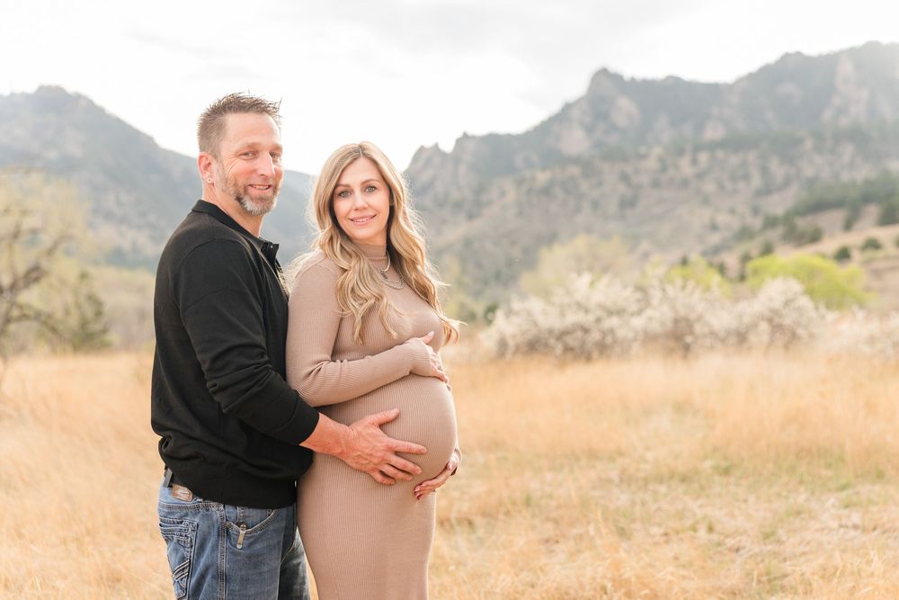 Couple posing in golden field with mountains, woman in beige maternity dress, man in black shirt embracing her baby bump.