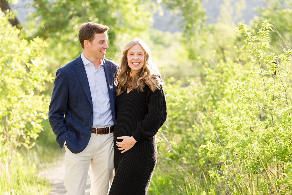 Happy couple smiling outdoors on a sunny path surrounded by lush green trees and bushes.