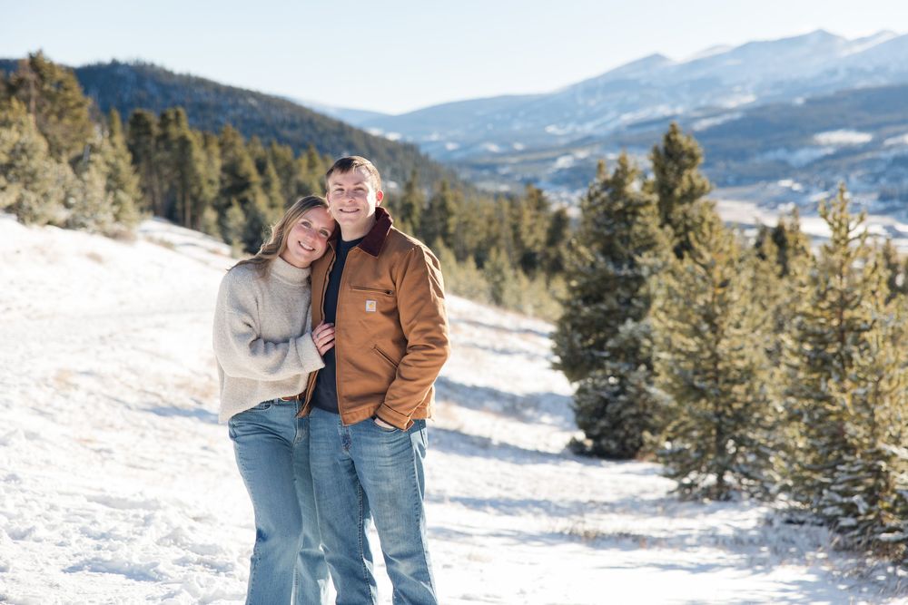 Couple embracing and smiling in a snowy mountain landscape with pine trees and blue skies in winter.
