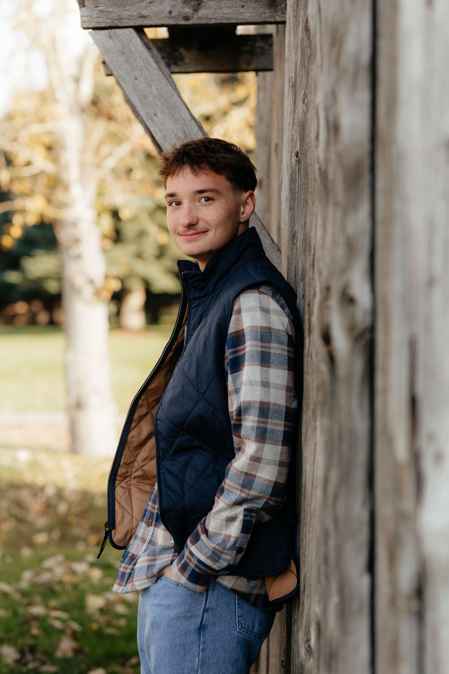 Young man in plaid shirt and navy vest leaning against rustic wooden barn in autumn setting.