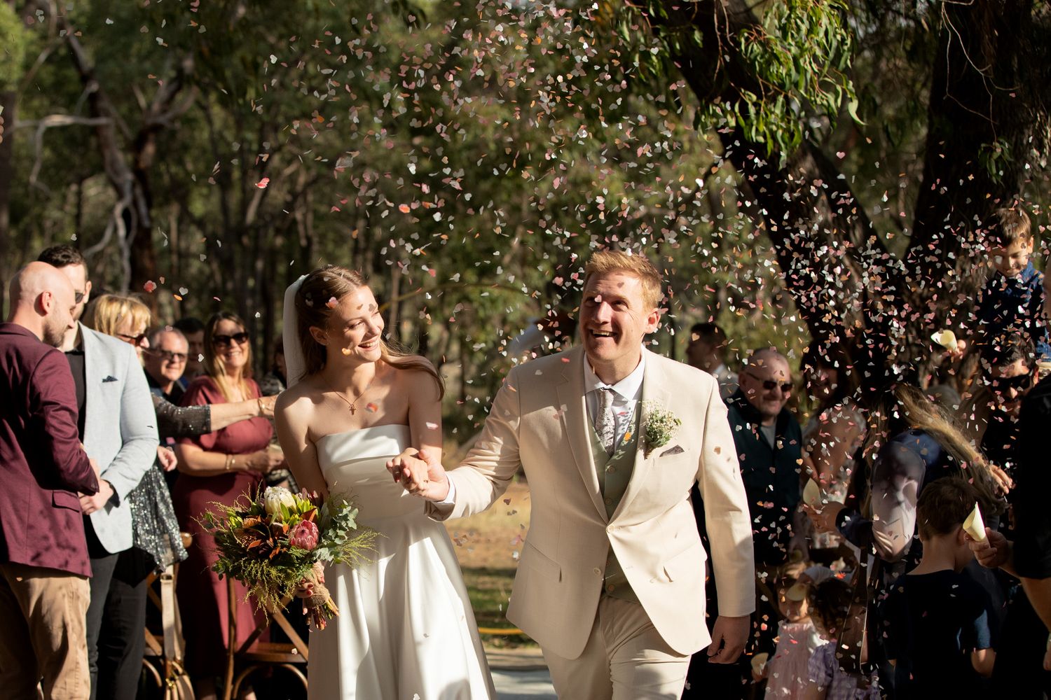 Joyful newlyweds walk through confetti shower surrounded by guests in a sunlit outdoor wedding ceremony.