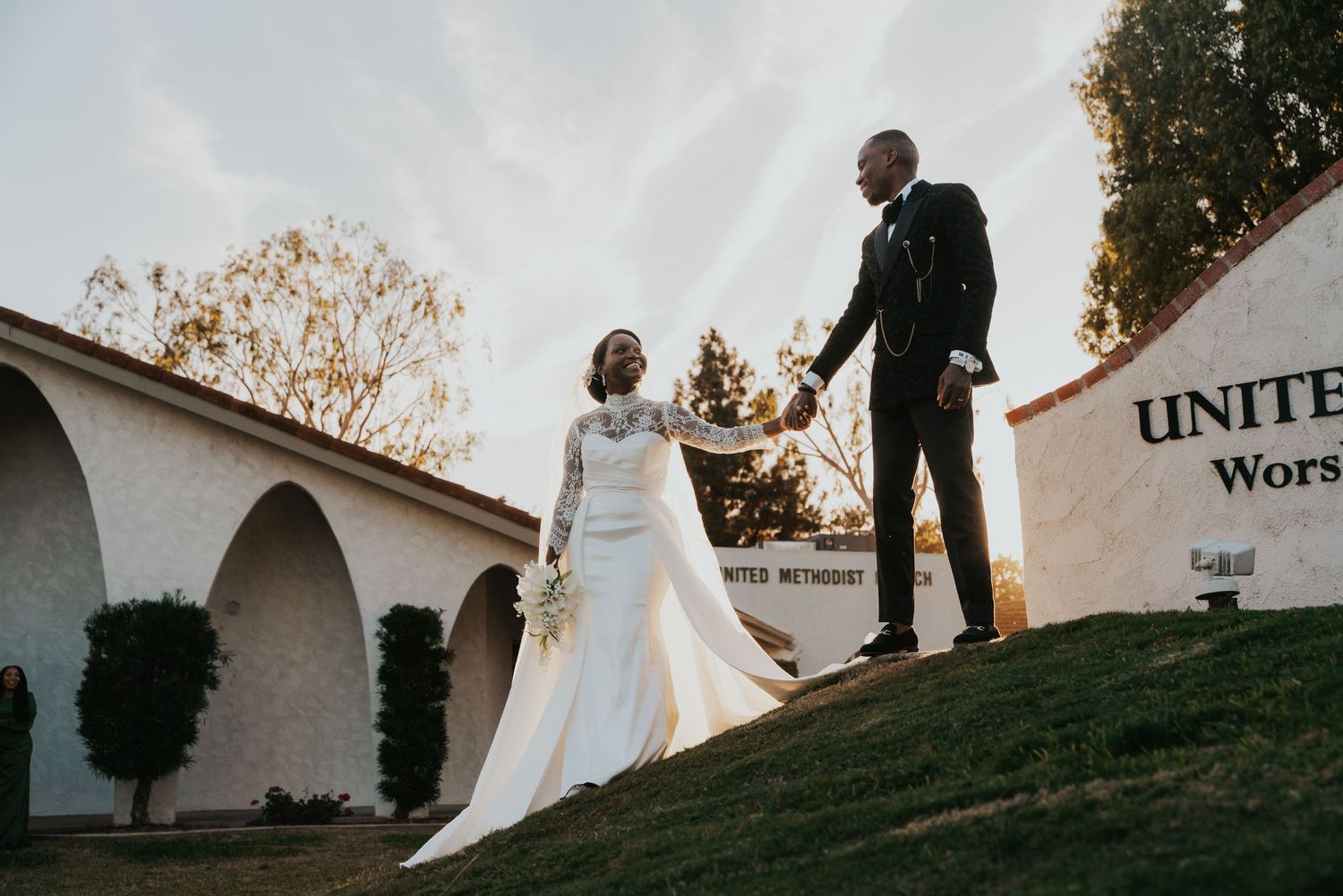 A couple in a white wedding dress and turquoise suit pose together on steps by a dark brick wall.