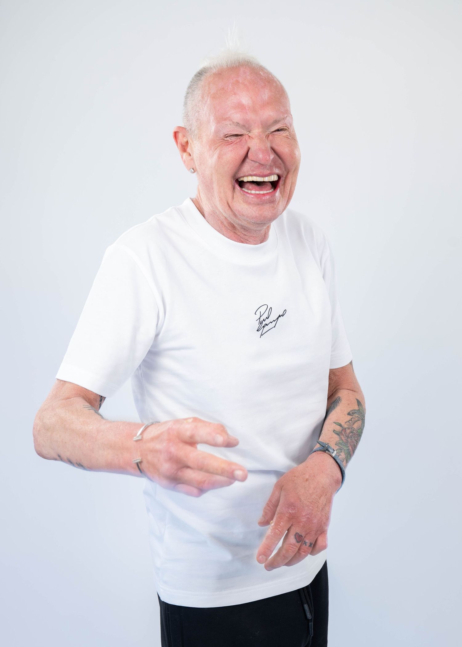 Elderly man with white hair laughing joyfully, wearing a white t-shirt with tattoos on his arms.