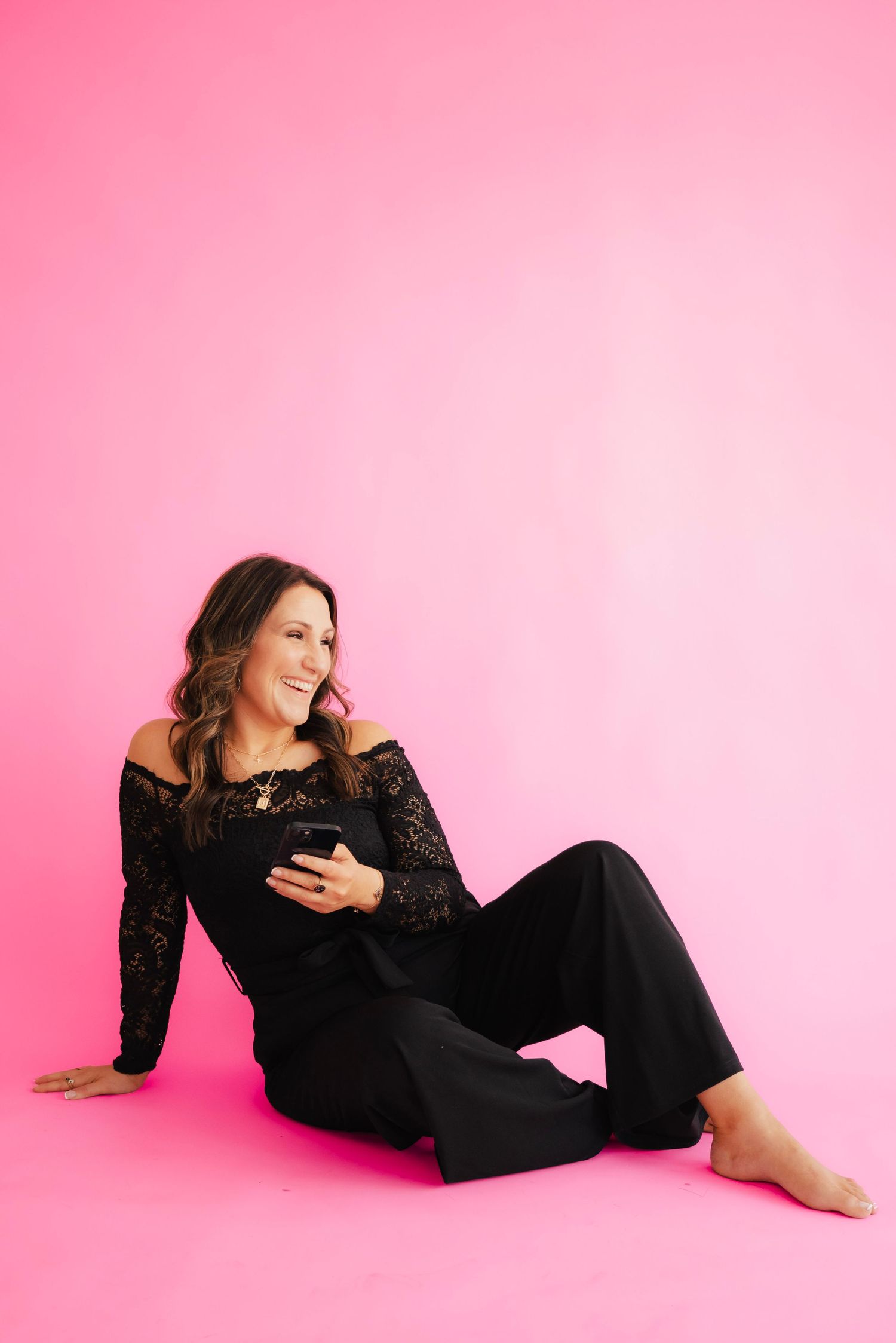 Woman in black lace off-shoulder top and pants sits on pink studio floor, smiling and holding a smartphone.