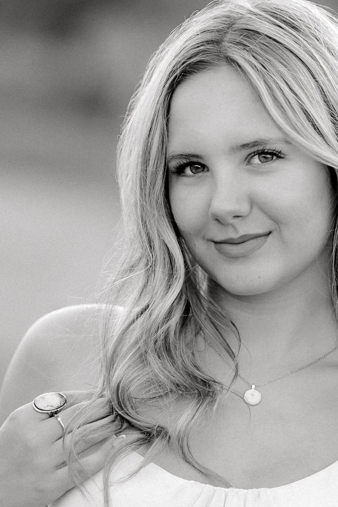 Black and white portrait of a young blonde woman wearing a necklace and ring, smiling softly outdoors.
