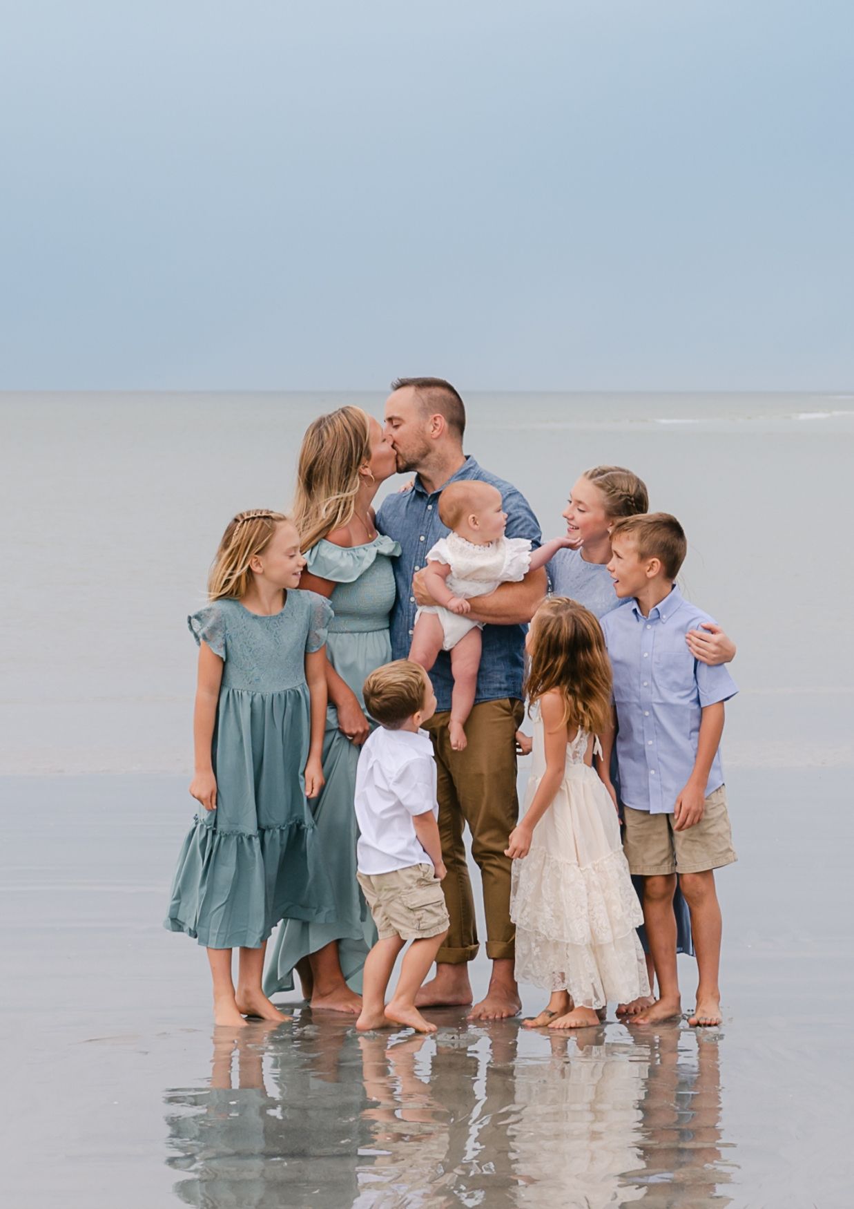 Family of seven posing together on a beach, parents kissing while children gather around them in coordinated outfits.