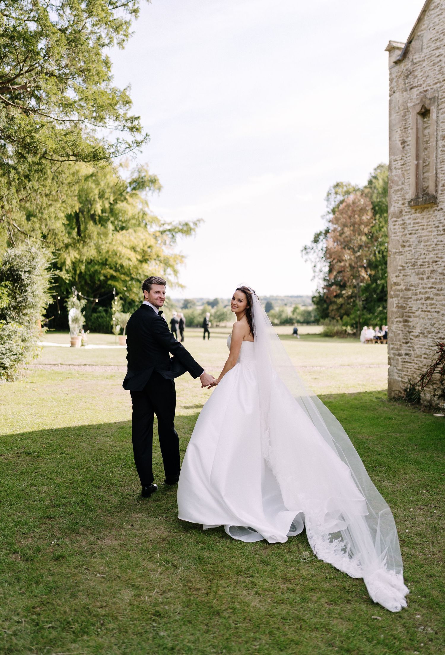 Bride in white gown with veil and groom in black tuxedo hold hands on green lawn beside stone building. Couple Shot's with Stu Tucker Photo