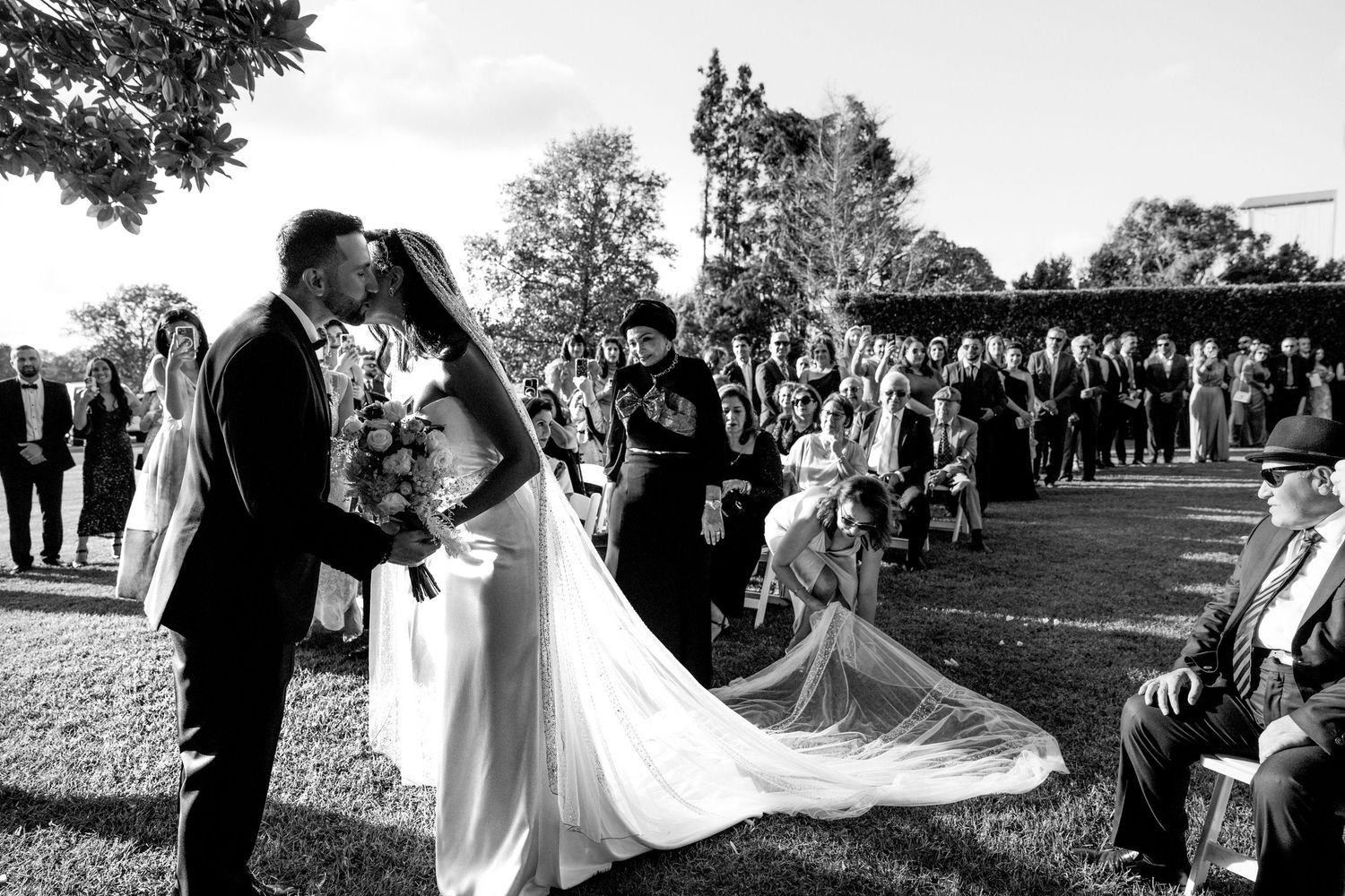 Black and white outdoor wedding ceremony photo of bride and groom kissing while guests celebrate on a sunny day.