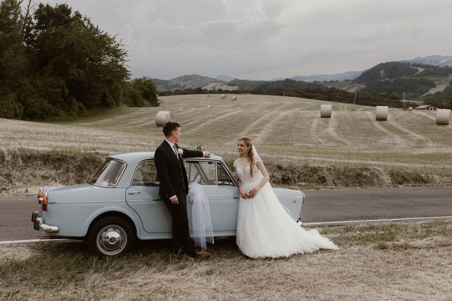 Bride and groom pose beside a vintage light blue car on a scenic rural road with hay bales and rolling hills.