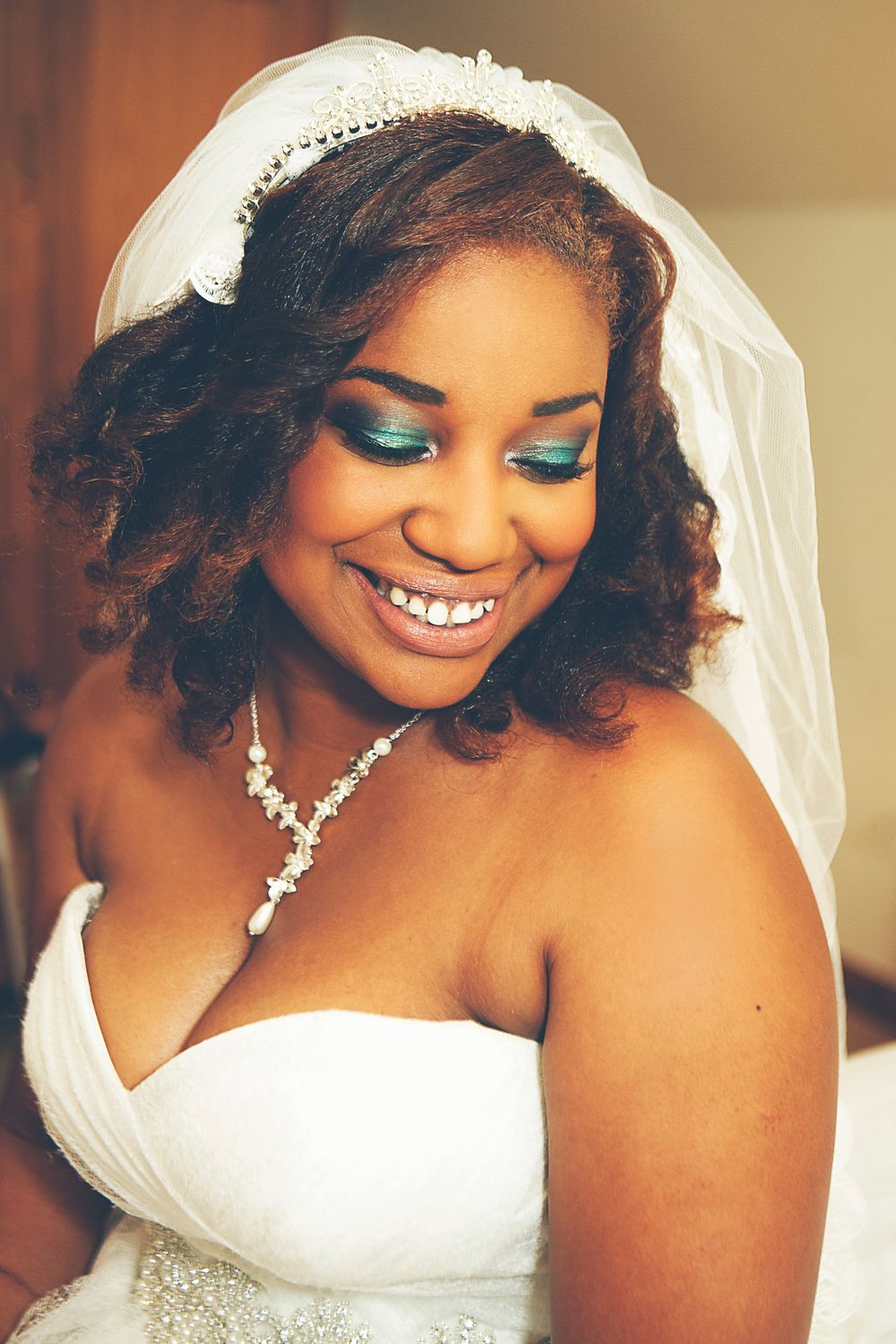 Smiling Black bride in white strapless gown, pearl necklace, tiara, and veil with teal eye makeup on wedding day. at Oaks farm in Surrey