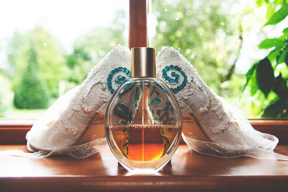 Elegant perfume bottle with ornate lace bridal shoes on a wooden windowsill with green foliage backdrop. Oaks farm wedding venue in Surrey
