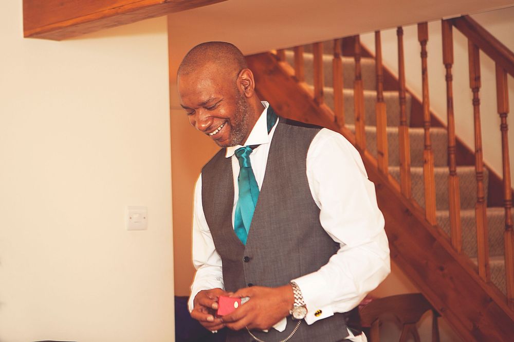 A smiling man in a grey vest and teal tie checks his phone near a wooden staircase. Oaks farm wedding venue in Surrey