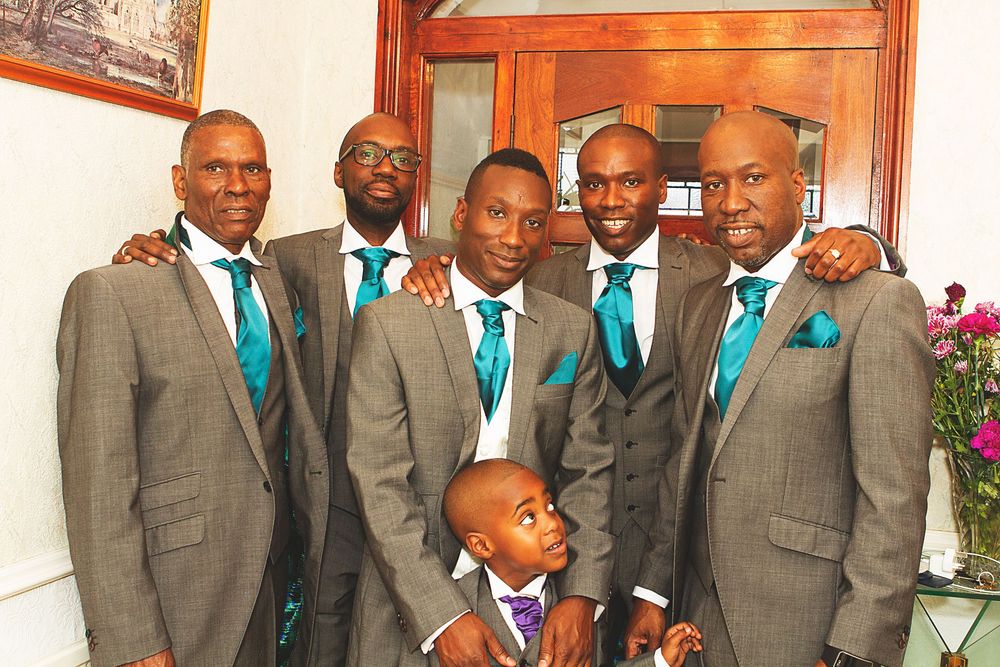 Six men in matching gray suits with teal ties pose together at a wedding, with a young boy in front. At Oaks farm wedding venue in Surrey