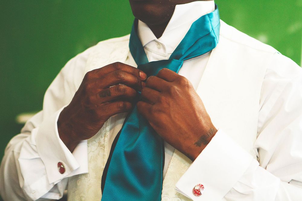 A man in a white dress shirt adjusts his teal tie, showcasing elegant cufflinks against a green background. At Oaks farm wedding venue.