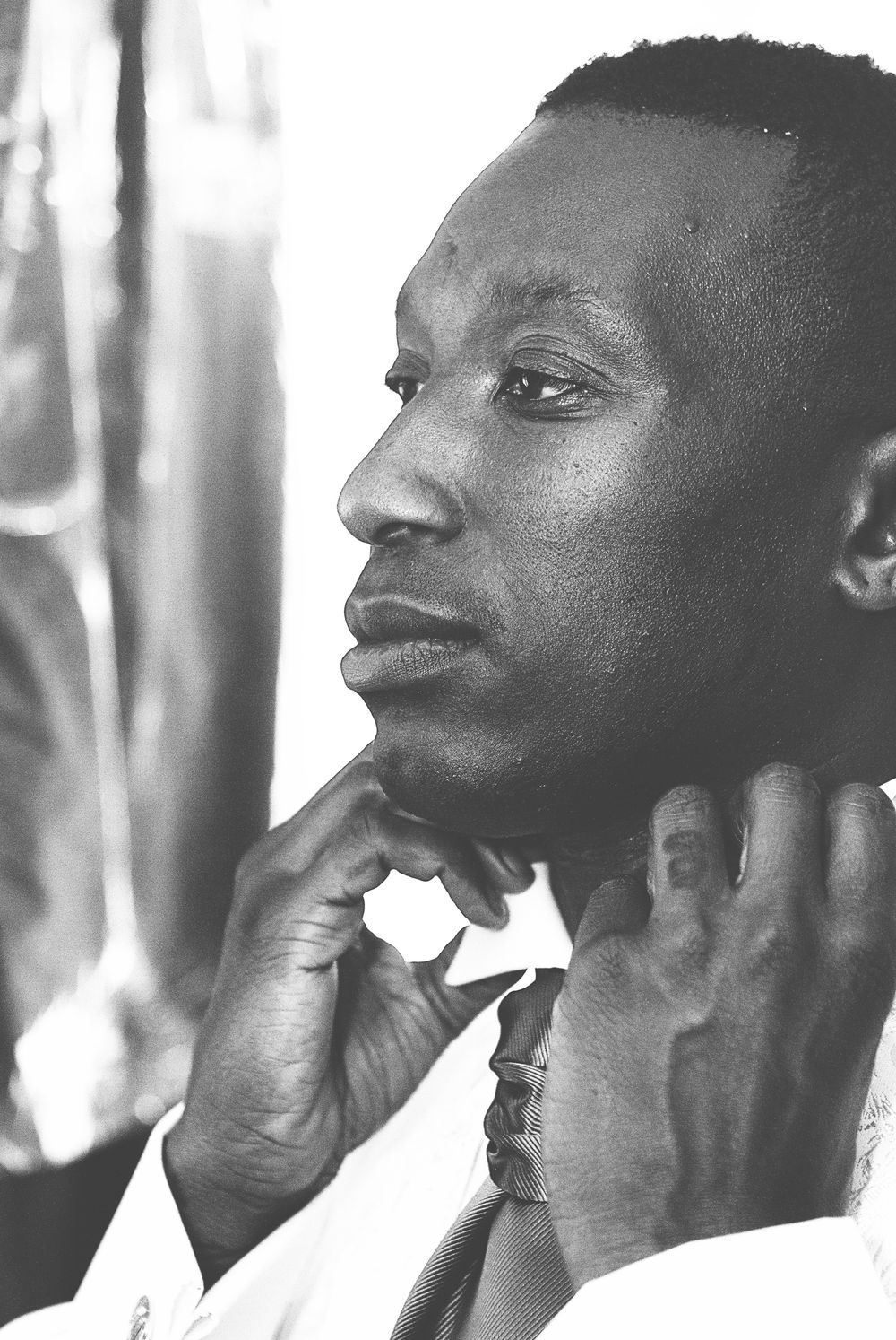 Black and white portrait of a man adjusting his tie, looking thoughtfully to the side. At Oaks farm wedding venue in Surrey