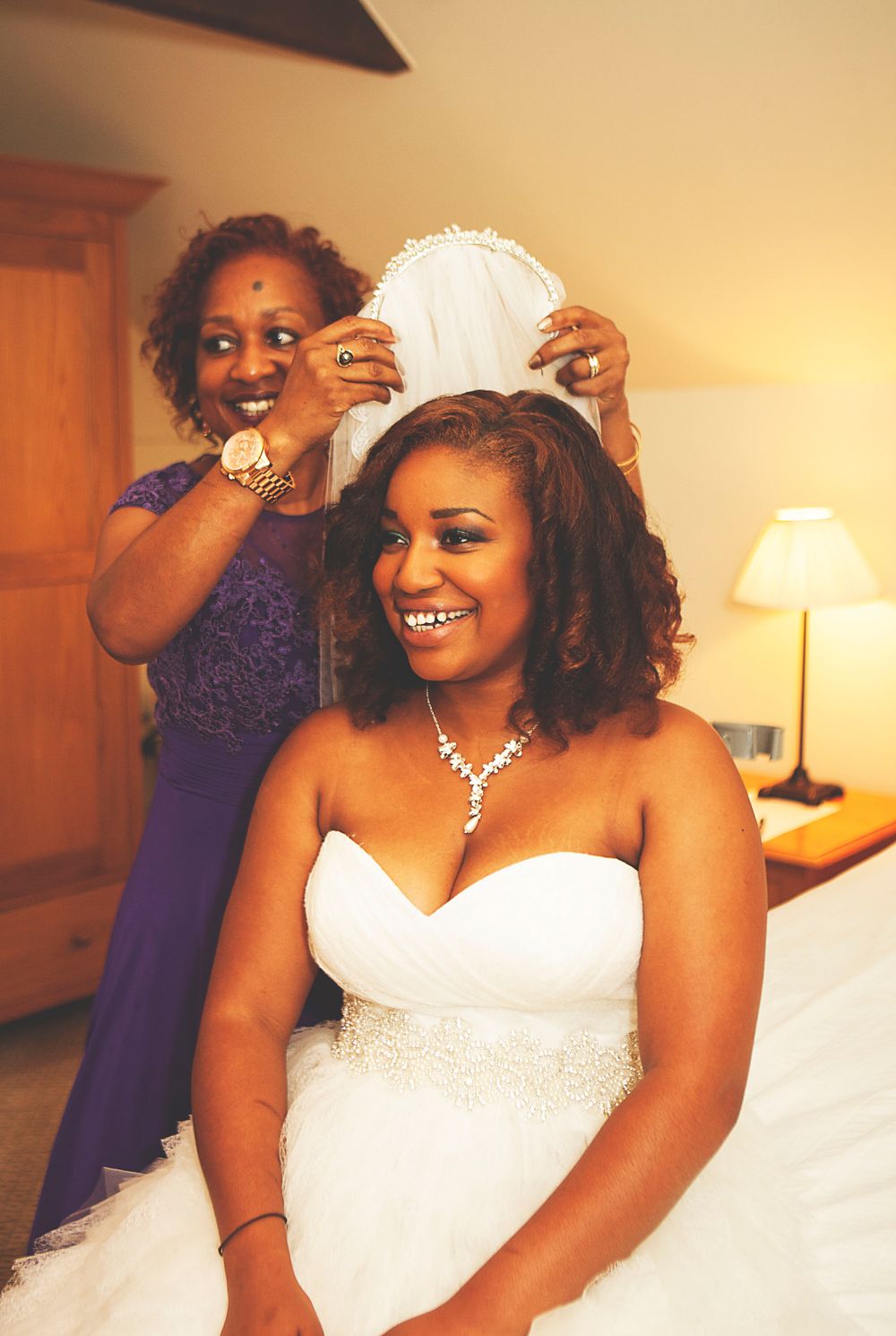 Bride in white strapless gown smiles as woman in purple dress places veil on her hair in hotel room. Oaks farm wedding venue in Surrey
