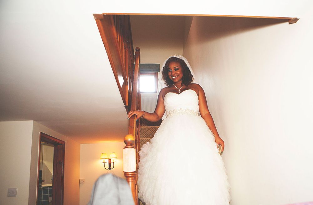 Bride in a strapless white ball gown and veil descends a wooden staircase, smiling radiantly on her wedding day. At Oaks farm wedding venue.