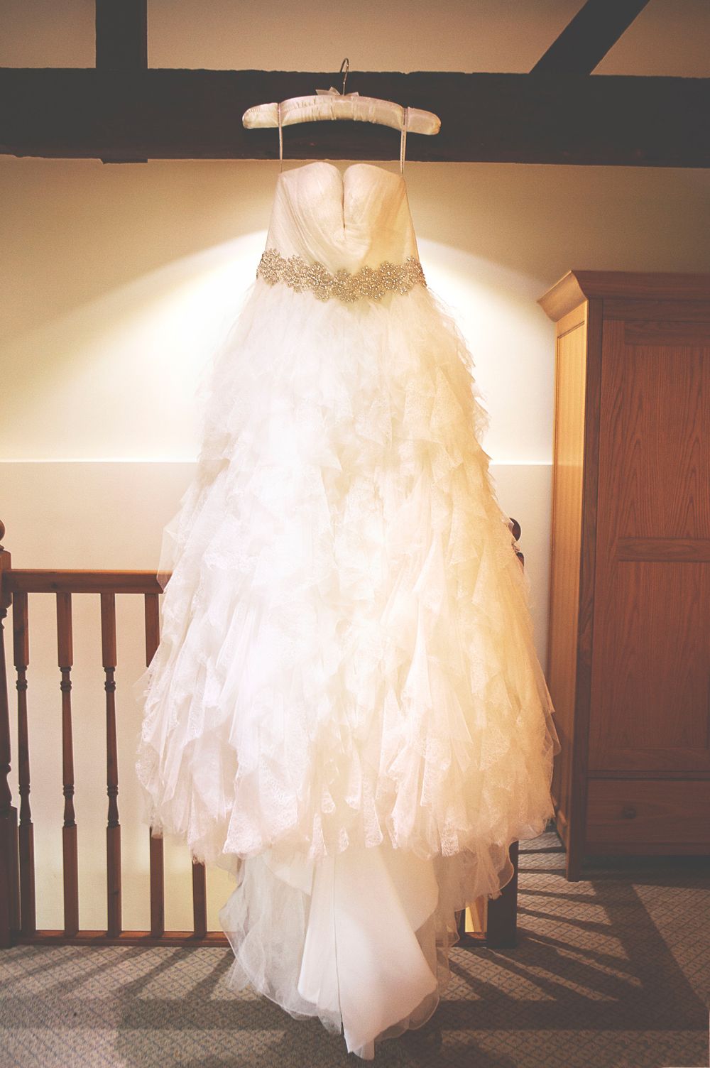 Elegant strapless white wedding dress with ruffled skirt and jeweled belt hanging on a hanger in a warmly lit room at Oaks farm.