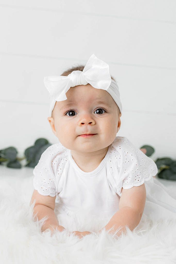 Adorable baby girl in white eyelet onesie and bow headband sitting on fluffy white fur with eucalyptus.