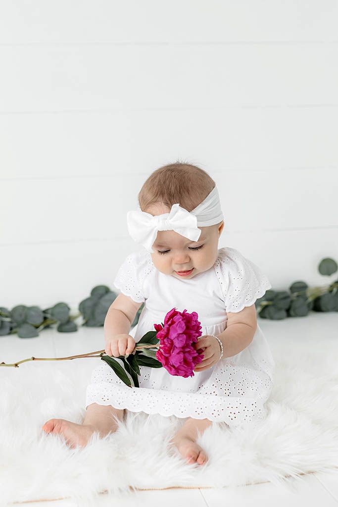 Adorable baby girl in white dress and bow headband sitting on fur rug holding a pink peony flower.