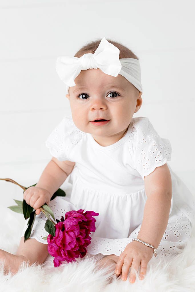 Adorable baby girl in white eyelet dress and bow headband holds pink peony flower on fluffy white fur.