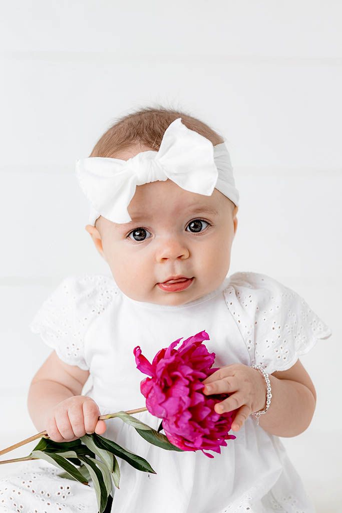 Adorable baby girl in white dress and bow headband holding a pink peony flower.