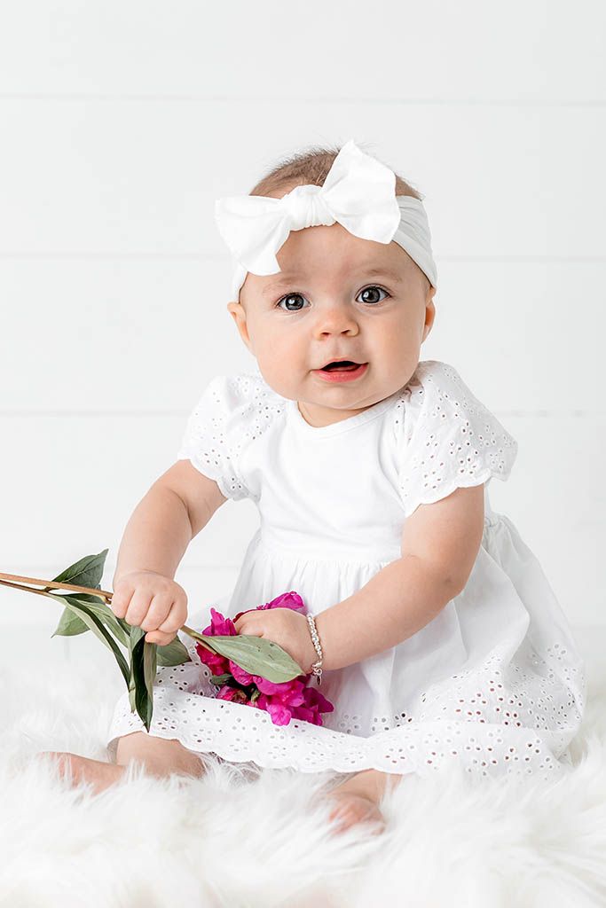 Cute baby girl in white eyelet dress holding pink peony flower on fluffy white rug.