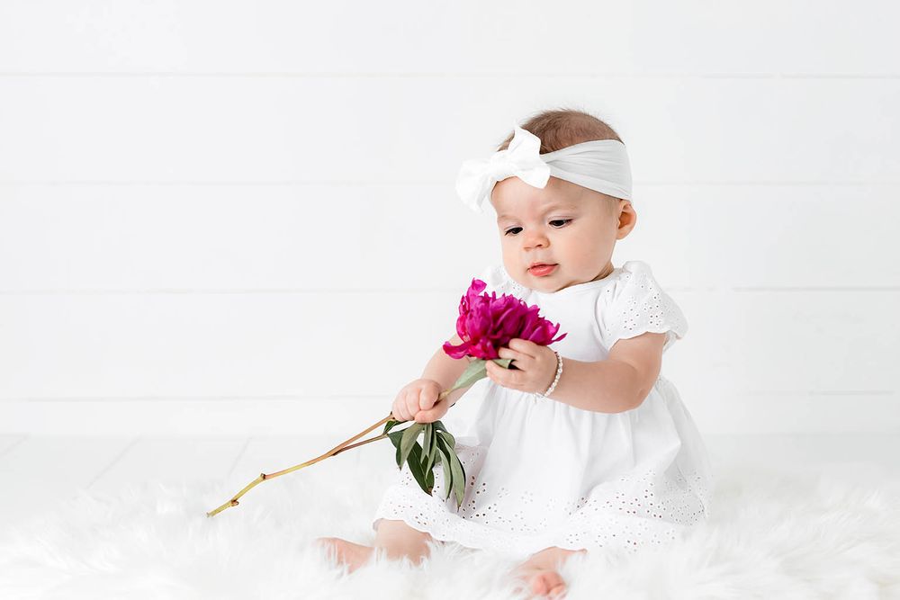 Sweet baby girl in white dress with bow headband examines a vibrant pink peony flower on white fur background.