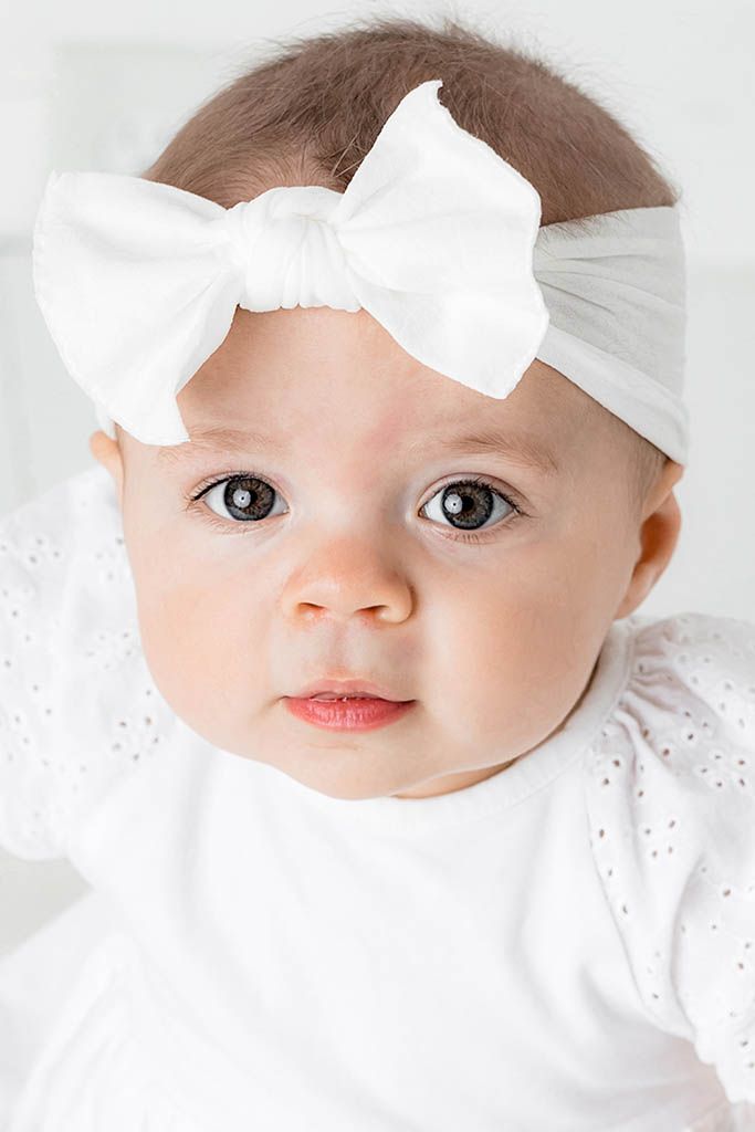 Close-up portrait of a cute baby girl wearing a white bow headband and white eyelet onesie.