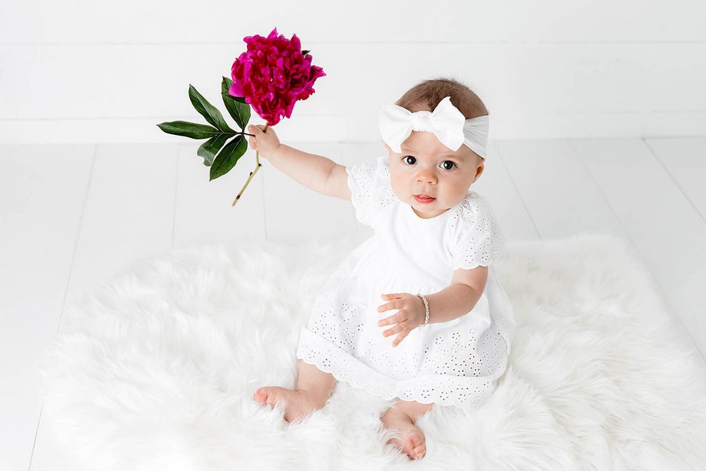 Sweet baby girl in white dress and bow headband holding up a bright pink peony flower.
