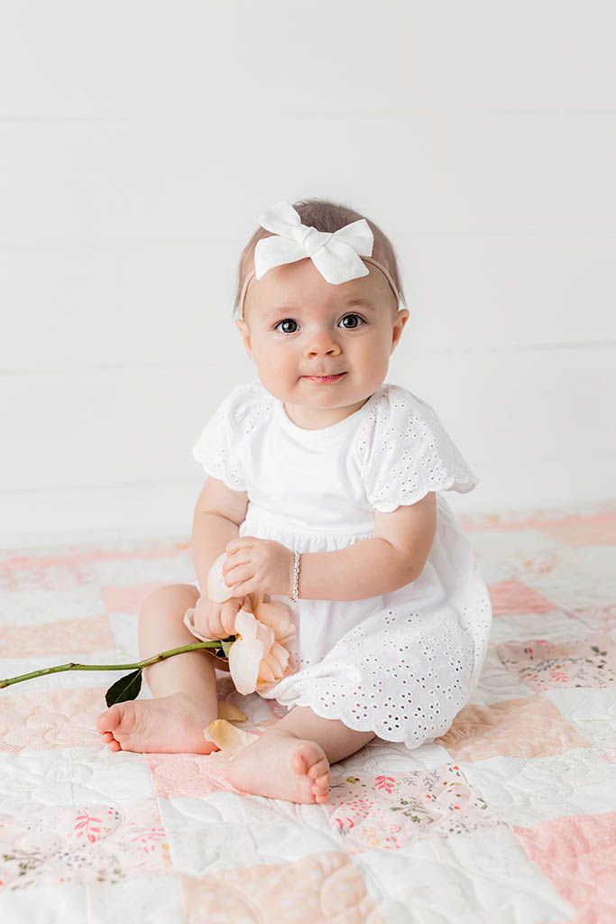 Baby girl in white eyelet dress and bow headband sits on floral quilt holding a white flower.